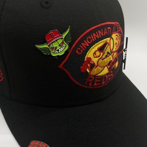 May include: Black baseball cap with a red and yellow embroidered Cincinnati Reds logo, a green goblin pin with a red hat, and a silver pin with the word "FADE" in red letters.