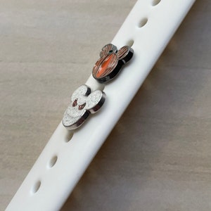 May include: Two small, silver-colored Mickey Mouse charms with enamel paint. One charm is orange and the other is white with glitter.