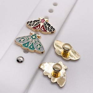 May include: Three gold-colored metal charms in the shape of moths. One moth is black with red and white accents, one is gray with green and white accents, and one is plain gold. The charms are on a white background.