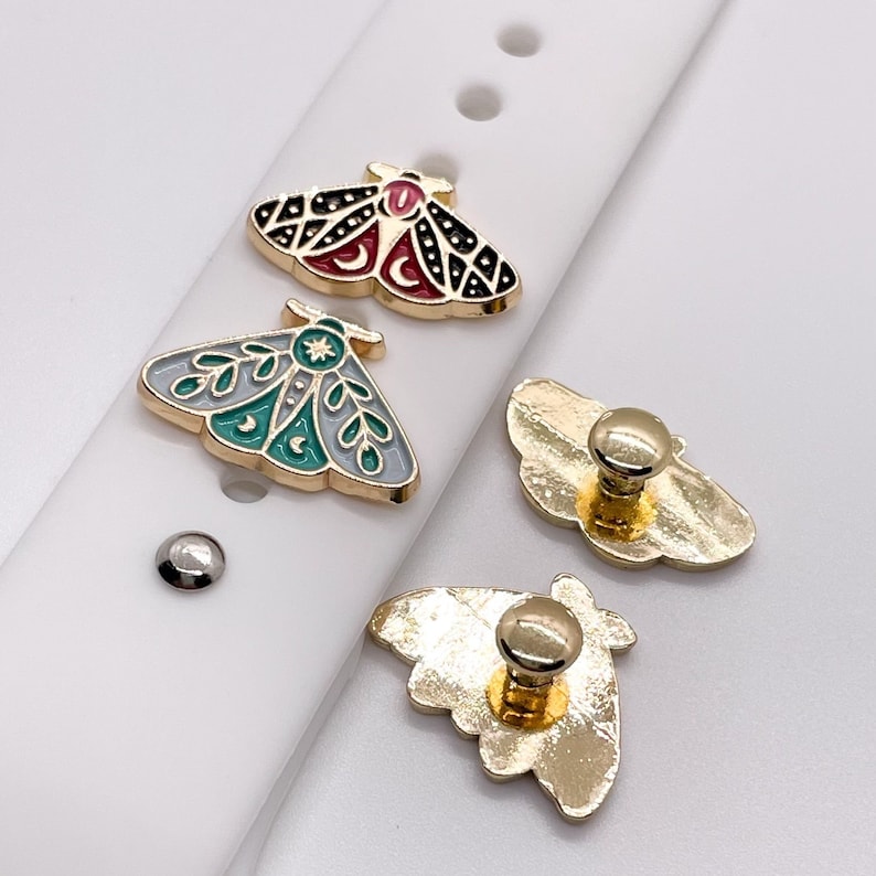 May include: Three gold-colored metal charms in the shape of moths. One moth is black with red and white accents, one is gray with green and white accents, and one is plain gold. The charms are on a white background.