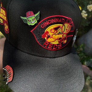 May include: Black baseball cap with a red and yellow Cincinnati Reds logo and a green goblin pin. The cap has a black brim and a red and yellow pin on the front.