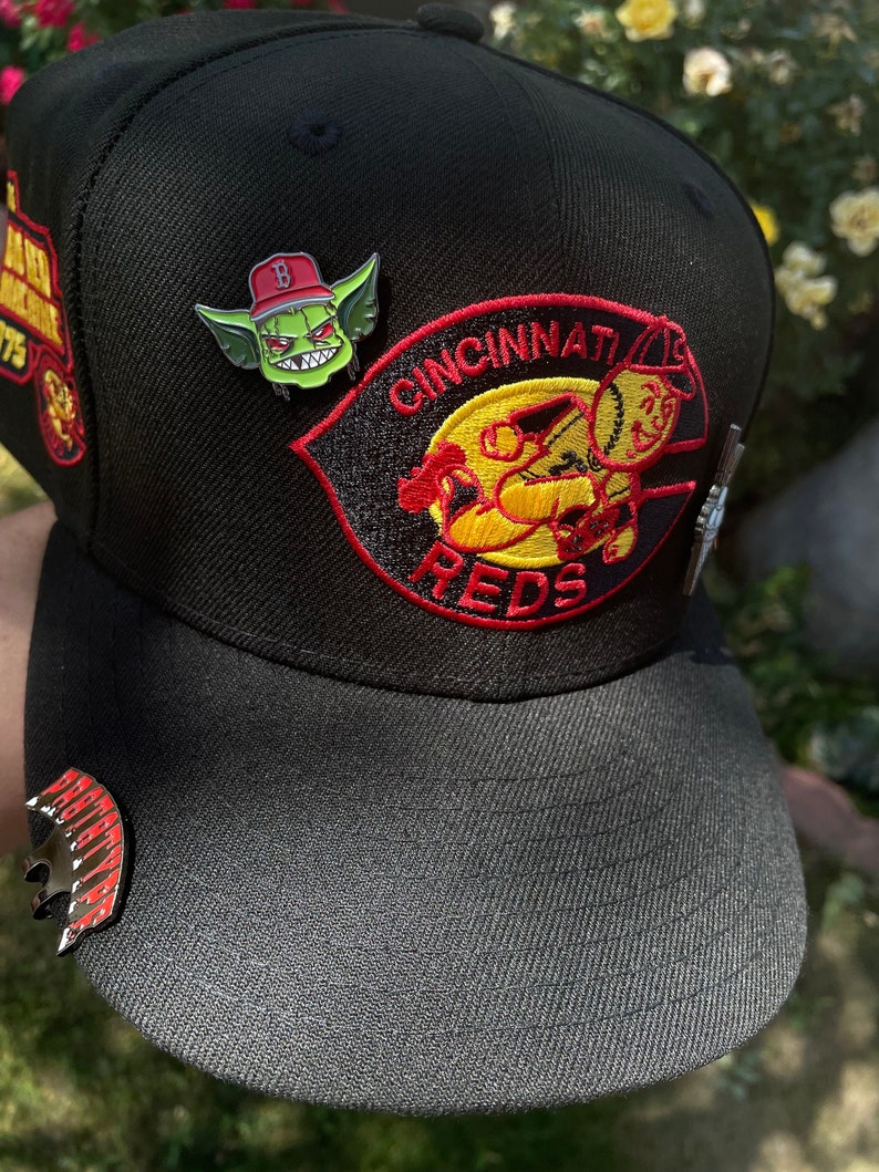 May include: Black baseball cap with a red and yellow Cincinnati Reds logo and a green goblin pin. The cap has a black brim and a red and yellow pin on the front.