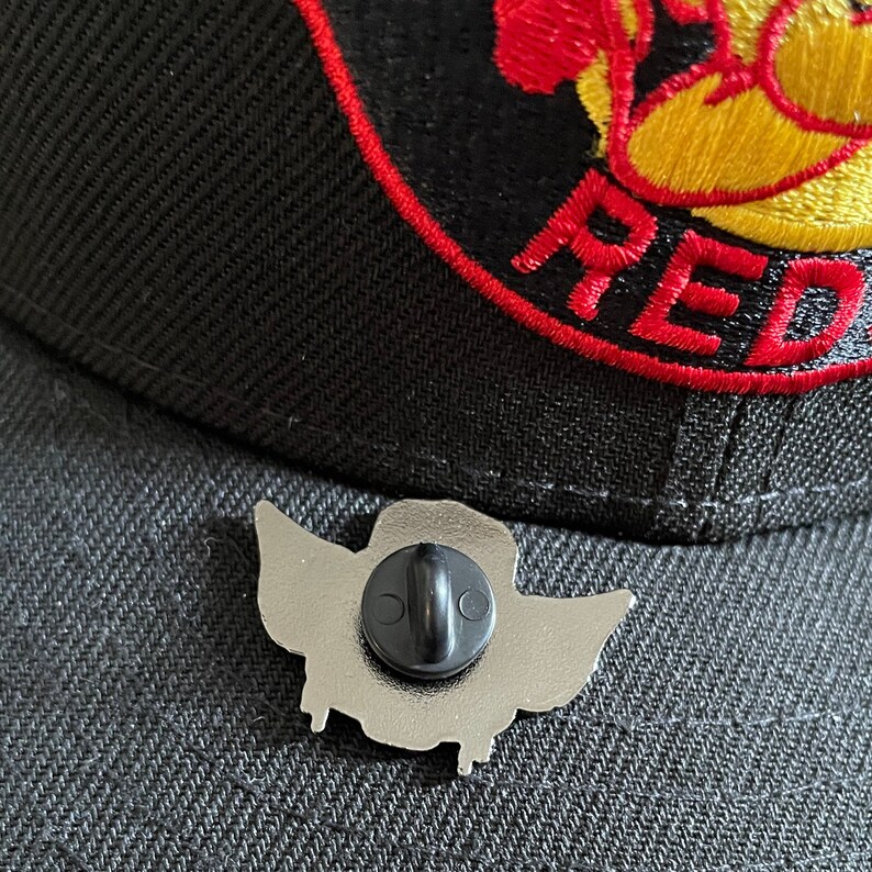 May include: Silver-toned enamel pin with a cartoon character wearing a hat and wings. The pin has a black rubber clutch back.