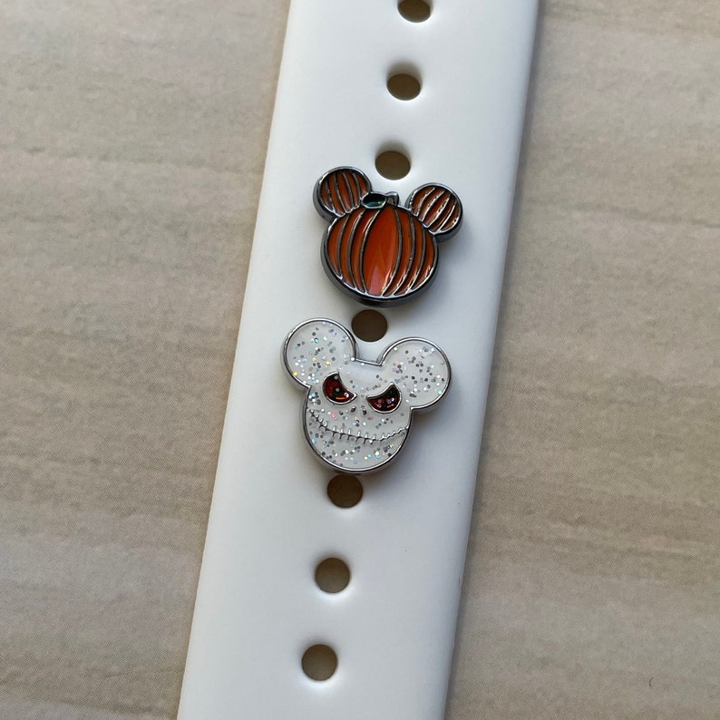 May include: Two Halloween-themed Mickey Mouse watch band charms. One charm is a pumpkin with a green stem and the other is a white Mickey Mouse head with a black and white Jack Skellington face.
