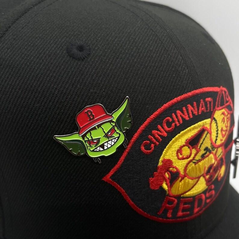 May include: Black baseball cap with a red and yellow embroidered "Cincinnati Reds" logo and a green and red goblin wearing a red baseball cap with a "B" on it.