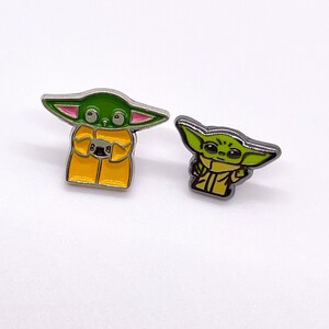 May include: Two enamel pins featuring a green alien character with large ears. The larger pin shows the character wearing a yellow raincoat and holding a small, round object. The smaller pin shows the character in a yellow shirt.