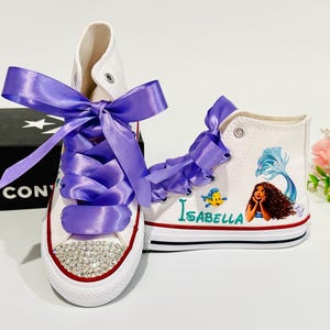 May include: White high-top sneakers with purple satin ribbon laces and a crystal-studded toe cap. One shoe features a mermaid graphic with the name "Isabella". A black Converse box is in the background.