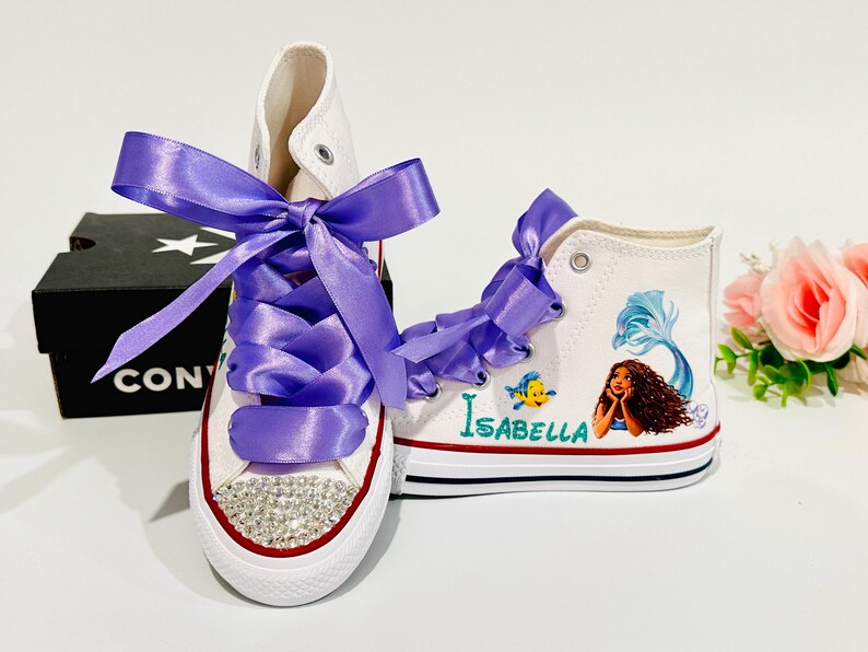 May include: White high-top sneakers with purple satin ribbon laces and a crystal-studded toe cap. One shoe features a mermaid graphic with the name "Isabella". A black Converse box is in the background.