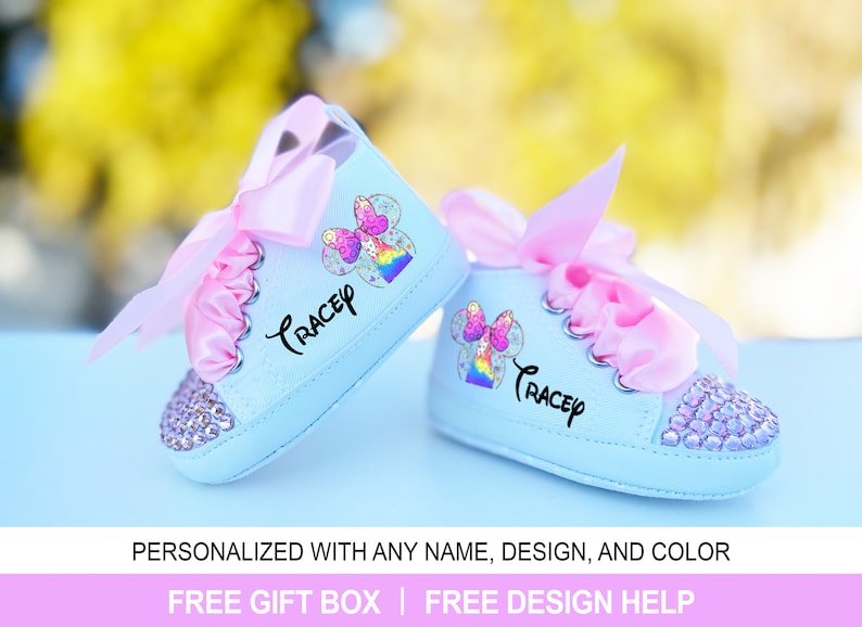 May include: A pair of light blue baby shoes with pink laces and a pink bow. The shoes are decorated with rhinestones and a colorful cartoon character with the name "Tracey" written on them.