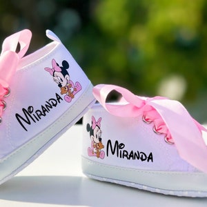 K&ouml;nnte beinhalten: Ein Paar wei&szlig;e Babyschuhe mit rosa Satinb&auml;ndern und einem Minnie Mouse-Motiv. Die Schuhe sind mit dem Namen "Miranda" personalisiert.