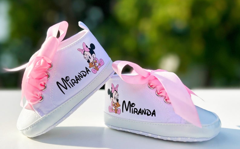 K&ouml;nnte beinhalten: Ein Paar wei&szlig;e Babyschuhe mit rosa Satinb&auml;ndern und einem Minnie Mouse-Motiv. Die Schuhe sind mit dem Namen "Miranda" personalisiert.