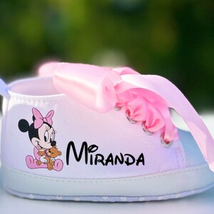 K&ouml;nnte beinhalten: Wei&szlig;e Babyschuhe mit rosa Schleifenb&auml;ndern und einem Minnie Mouse-Motiv. Die Schuhe sind mit dem Namen "Miranda" personalisiert.