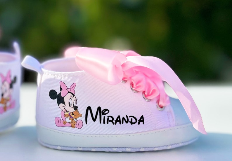 K&ouml;nnte beinhalten: Wei&szlig;e Babyschuhe mit rosa Schleifenb&auml;ndern und einem Minnie Mouse-Motiv. Die Schuhe sind mit dem Namen "Miranda" personalisiert.