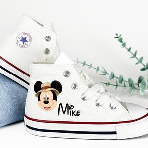 May include: White high-top sneakers with a red and navy stripe on the sole. One shoe features a Mickey Mouse graphic wearing a safari hat and the name "Mike". The other shoe has a Converse All Star logo.