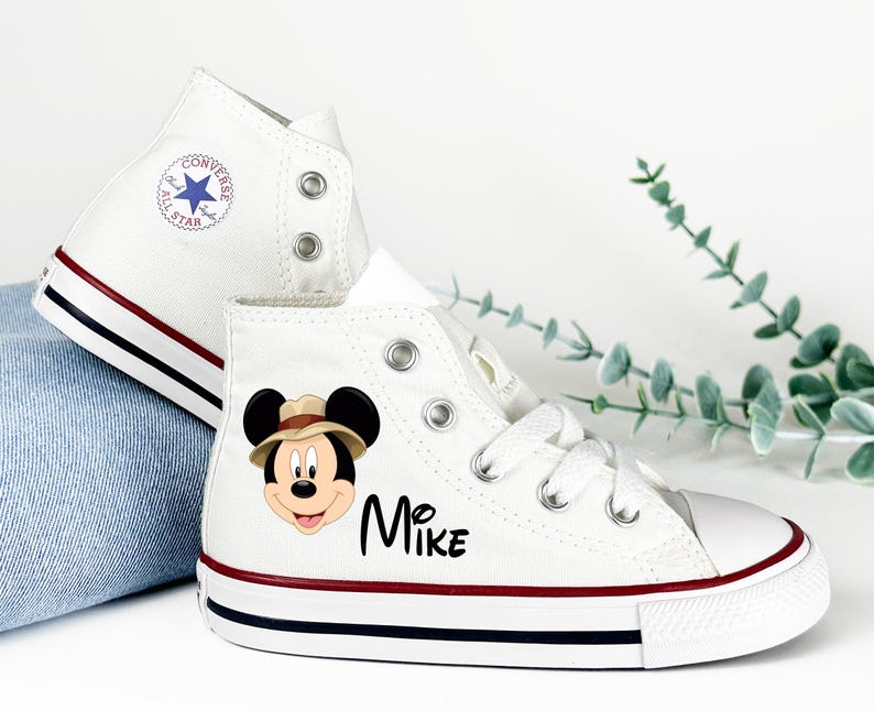 May include: White high-top sneakers with a red and navy stripe on the sole. One shoe features a Mickey Mouse graphic wearing a safari hat and the name "Mike". The other shoe has a Converse All Star logo.