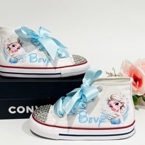 May include: White high-top sneakers with light blue ribbon laces and a rhinestone-studded toe cap. The shoes feature a cartoon image of a princess and the name "Bev" in light blue. The shoes are on a black box.