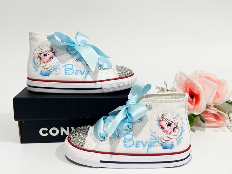 May include: White high-top sneakers with light blue ribbon laces and a rhinestone-studded toe cap. The shoes feature a cartoon image of a princess and the name "Bev" in light blue. The shoes are on a black box.