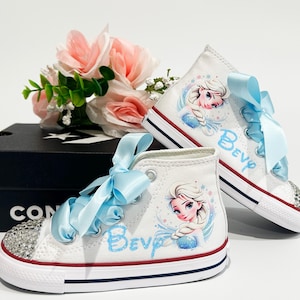 May include: White high-top sneakers with a cartoon image of a princess and the name "Bev" in blue script. The sneakers have light blue ribbon laces and rhinestone-studded toes. A black Converse box and pink flowers are in the background.