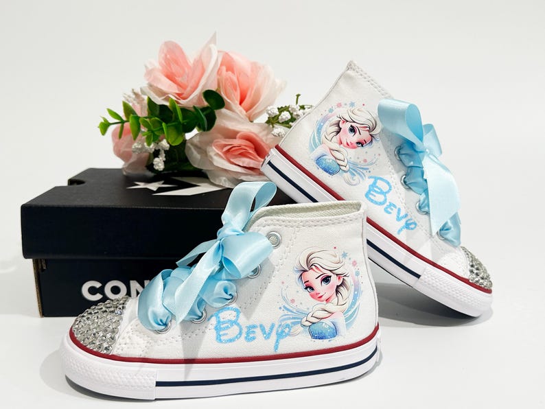 May include: White high-top sneakers with a cartoon image of a princess and the name "Bev" in blue script. The sneakers have light blue ribbon laces and rhinestone-studded toes. A black Converse box and pink flowers are in the background.