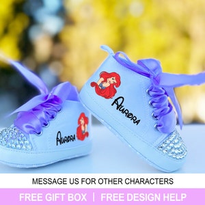May include: A pair of baby sneakers in light blue with a rhinestone-studded toe cap. Each shoe features a cartoon image of a mermaid and the name "Aurora" in black. The sneakers have purple ribbon laces.