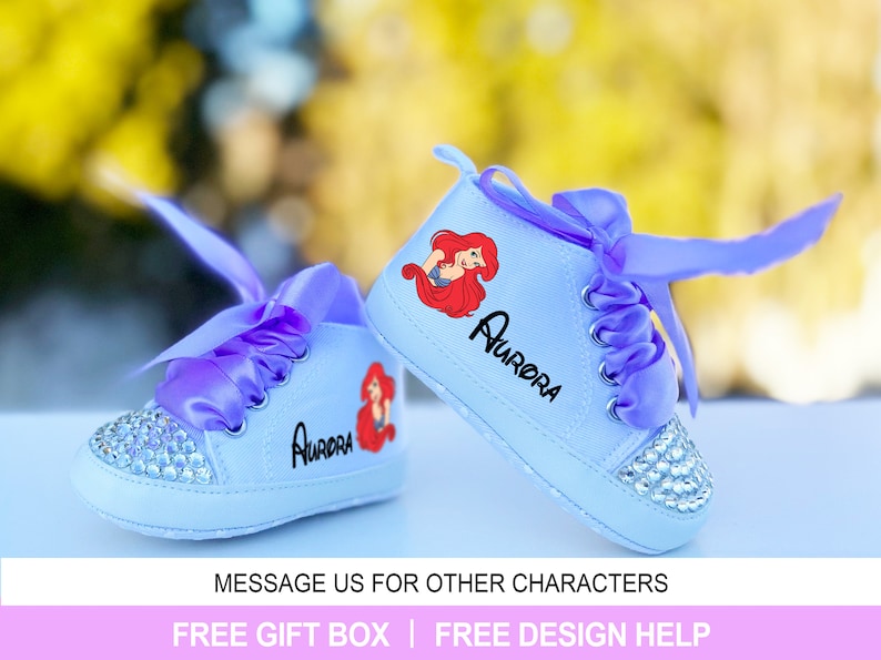 May include: A pair of baby sneakers in light blue with a rhinestone-studded toe cap. Each shoe features a cartoon image of a mermaid and the name "Aurora" in black. The sneakers have purple ribbon laces.