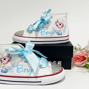 May include: White high-top sneakers with light blue ribbon laces and rhinestone-studded toes. The shoes feature a cartoon image of Elsa from Frozen and the name "Bev" in light blue script. Red and blue accents.
