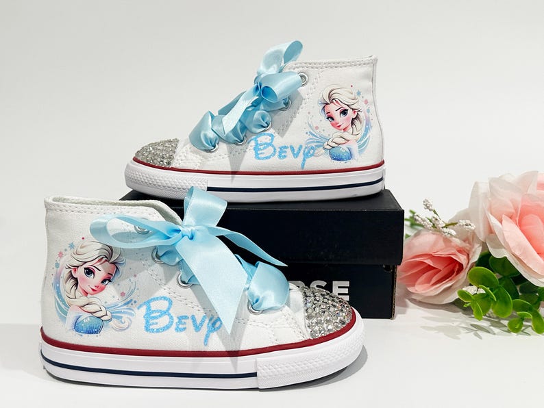 May include: White high-top sneakers with light blue ribbon laces and rhinestone-studded toes. The shoes feature a cartoon image of Elsa from Frozen and the name "Bev" in light blue script. Red and blue accents.