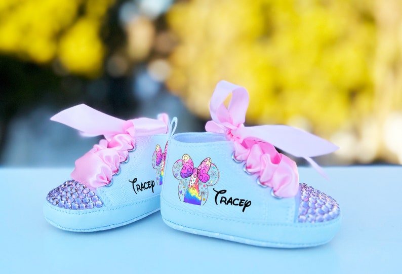 May include: A pair of light blue baby shoes with pink satin bows and rhinestones. The shoes have a rainbow and Mickey Mouse design with the name "Tracey" written on them.