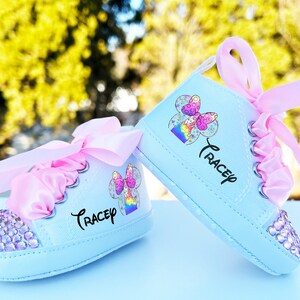 May include: A pair of light blue baby shoes with pink satin bows and rhinestones. The shoes have a colorful cartoon character design and the name "Trace" written on them.