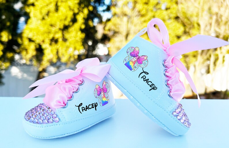 May include: A pair of light blue baby shoes with pink satin bows and rhinestones. The shoes have a colorful cartoon character design and the name "Trace" written on them.