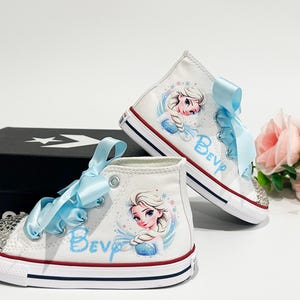 May include: White high-top sneakers with a cartoon image of a princess, light blue ribbon laces, and rhinestone accents. The word "Bev" is written in light blue on the side of the shoe. The shoes are sitting next to a black box and pink flowers.