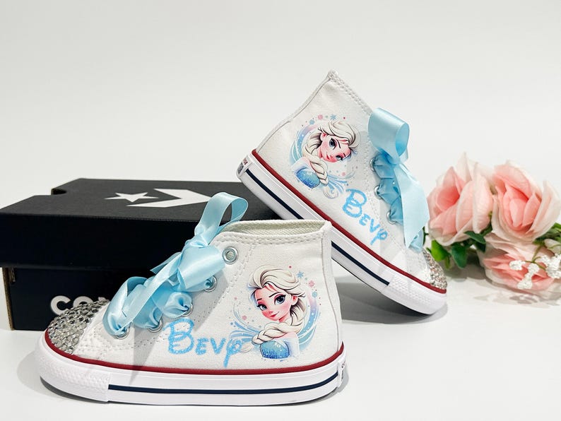 May include: White high-top sneakers with a cartoon image of a princess, light blue ribbon laces, and rhinestone accents. The word "Bev" is written in light blue on the side of the shoe. The shoes are sitting next to a black box and pink flowers.