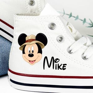 May include: White high-top canvas sneakers featuring a Mickey Mouse graphic wearing a safari hat. The name "Mike" is printed in black script on the side. The sneakers have red and blue stripes on the sole.