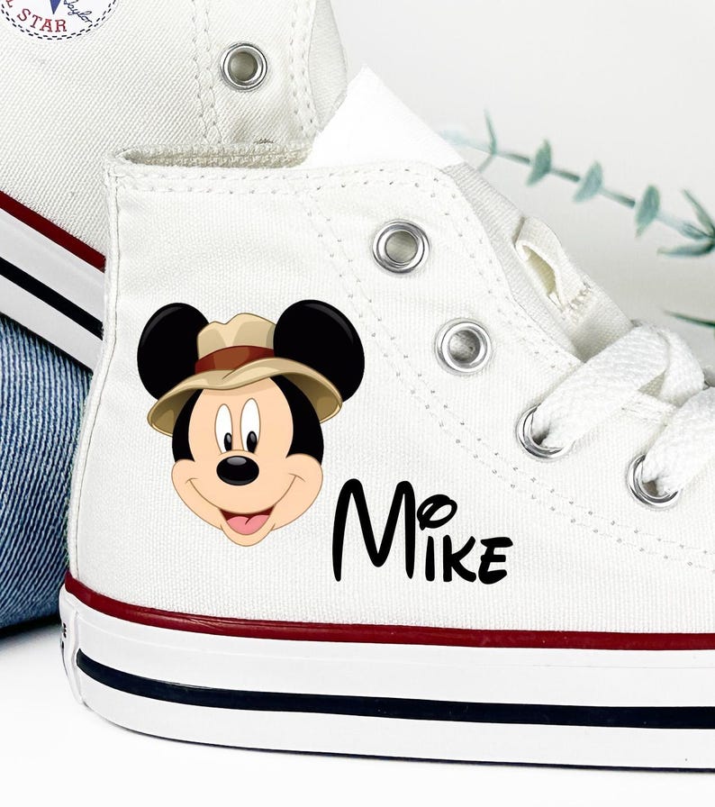 May include: White high-top canvas sneakers featuring a Mickey Mouse graphic wearing a safari hat. The name "Mike" is printed in black script on the side. The sneakers have red and blue stripes on the sole.