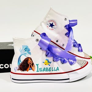 May include: White high-top sneakers with a mermaid design, purple ribbon laces, and the name "Isabella". The shoes feature a red and blue stripe on the sole and a sparkling toe cap. A black shoe box is in the background.