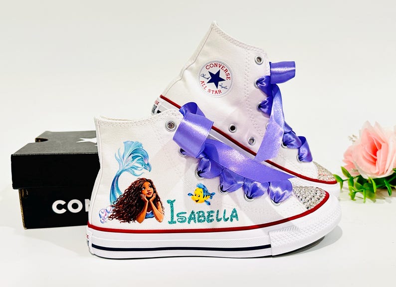 May include: White high-top sneakers with a mermaid design, purple ribbon laces, and the name "Isabella". The shoes feature a red and blue stripe on the sole and a sparkling toe cap. A black shoe box is in the background.