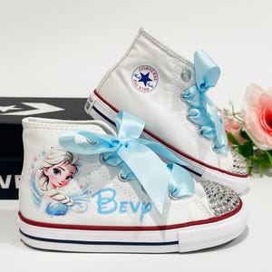 May include: White high-top Converse sneakers with light blue satin ribbon laces. One shoe features a rhinestone-studded toe cap and a cartoon image of Elsa from Frozen. The other shoe has the Converse logo.