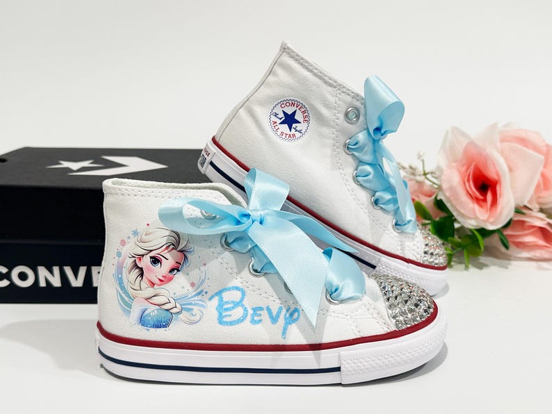 May include: White high-top Converse sneakers with light blue satin ribbon laces. One shoe features a rhinestone-studded toe cap and a cartoon image of Elsa from Frozen. The other shoe has the Converse logo.