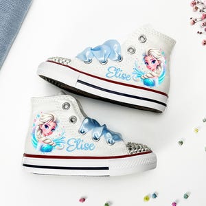 May include: White high-top sneakers with blue ribbon laces and a rhinestone-studded toe cap. Each shoe features a colorful illustration of a princess and the name "Elise" in blue script. The shoes have a red and blue stripe on the sole.