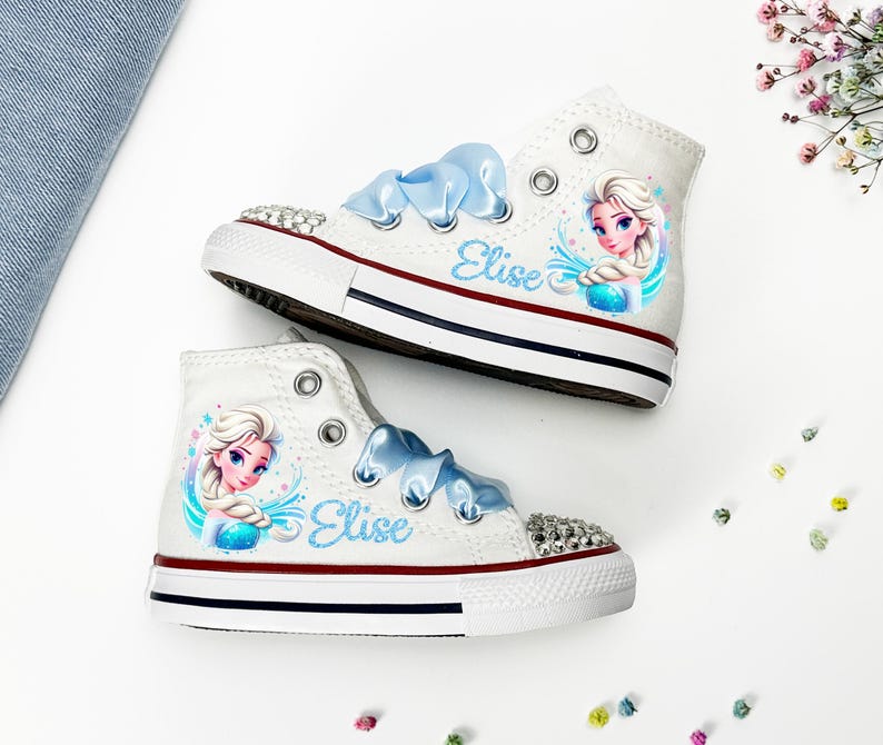 May include: White high-top sneakers with blue ribbon laces and a rhinestone-studded toe cap. Each shoe features a colorful illustration of a princess and the name "Elise" in blue script. The shoes have a red and blue stripe on the sole.