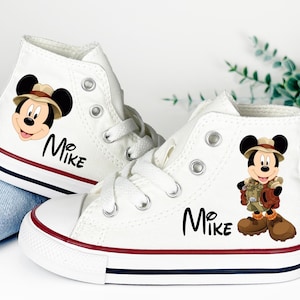 May include: White high-top canvas sneakers featuring a Mickey Mouse graphic and the name "Mike" in black script. The shoes have red and navy blue stripes on the soles. The Mickey Mouse graphic is wearing a safari outfit.