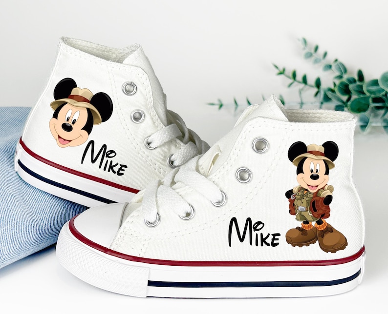 May include: White high-top canvas sneakers featuring a Mickey Mouse graphic and the name "Mike" in black script. The shoes have red and navy blue stripes on the soles. The Mickey Mouse graphic is wearing a safari outfit.