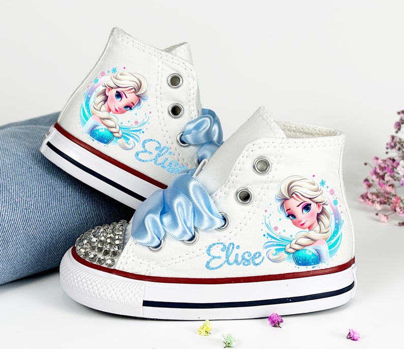 May include: White high-top sneakers featuring a cartoon image of a princess, with blue accents and ribbon laces. The toe cap is embellished with rhinestones. The word "Elise" is written in blue script on the side.