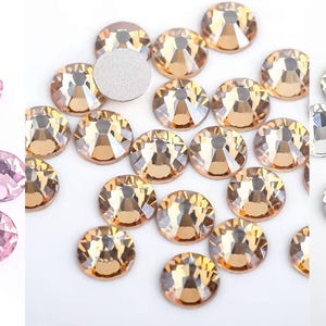 May include: A collection of round, faceted rhinestones in three colors: light pink, golden amber, and clear crystal. Each rhinestone has a flat, silver backing. The gems are scattered, showcasing their sparkle and clarity, ideal for craft projects.