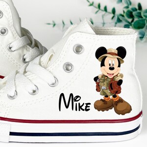 May include: White high-top canvas sneakers with red and blue trim. Features a cartoon graphic of Mickey Mouse in safari attire. The name "Mike" is printed in black script on the side.