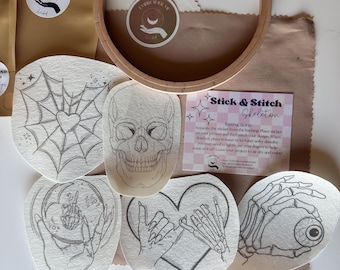 May include: A collection of embroidery supplies featuring a wooden hoop, fabric, and several Stick & Stitch designs. Designs include a spiderweb heart, skull, hands, and an eye. The packaging includes the text "EMBROIDER ME" and "Stick & Stitch Skeleton".