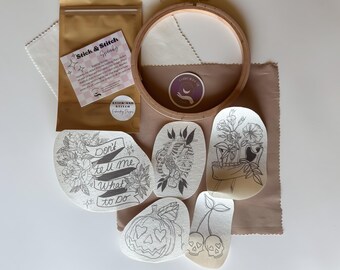 May include: Embroidery kit with a wooden hoop, fabric, and several pre-printed designs. Designs include floral arrangements, a skeleton, a pumpkin, and cherries with skulls. The kit also includes a gold-colored pouch with a sticker that reads "Stick & Stitch".