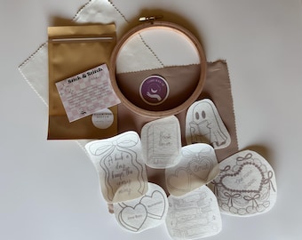 May include: A craft kit with a wooden embroidery hoop, fabric, and several sticker-like designs. The designs include a ghost reading a book, hearts, and text that reads "Book lover" and "One more chapter". A brown paper bag is also included.