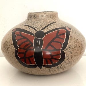 May include: A speckled, rounded vase with a large butterfly design. The butterfly has red wings with black outlines and a black body. The vase is a neutral tan color and sits on a white surface.