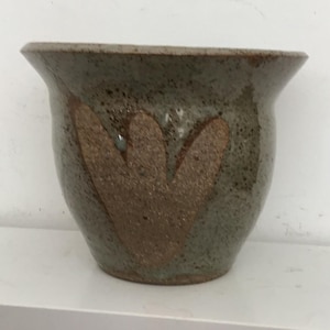 May include: A small, green ceramic planter with a brown, heart-shaped design. The planter is sitting on a white shelf.